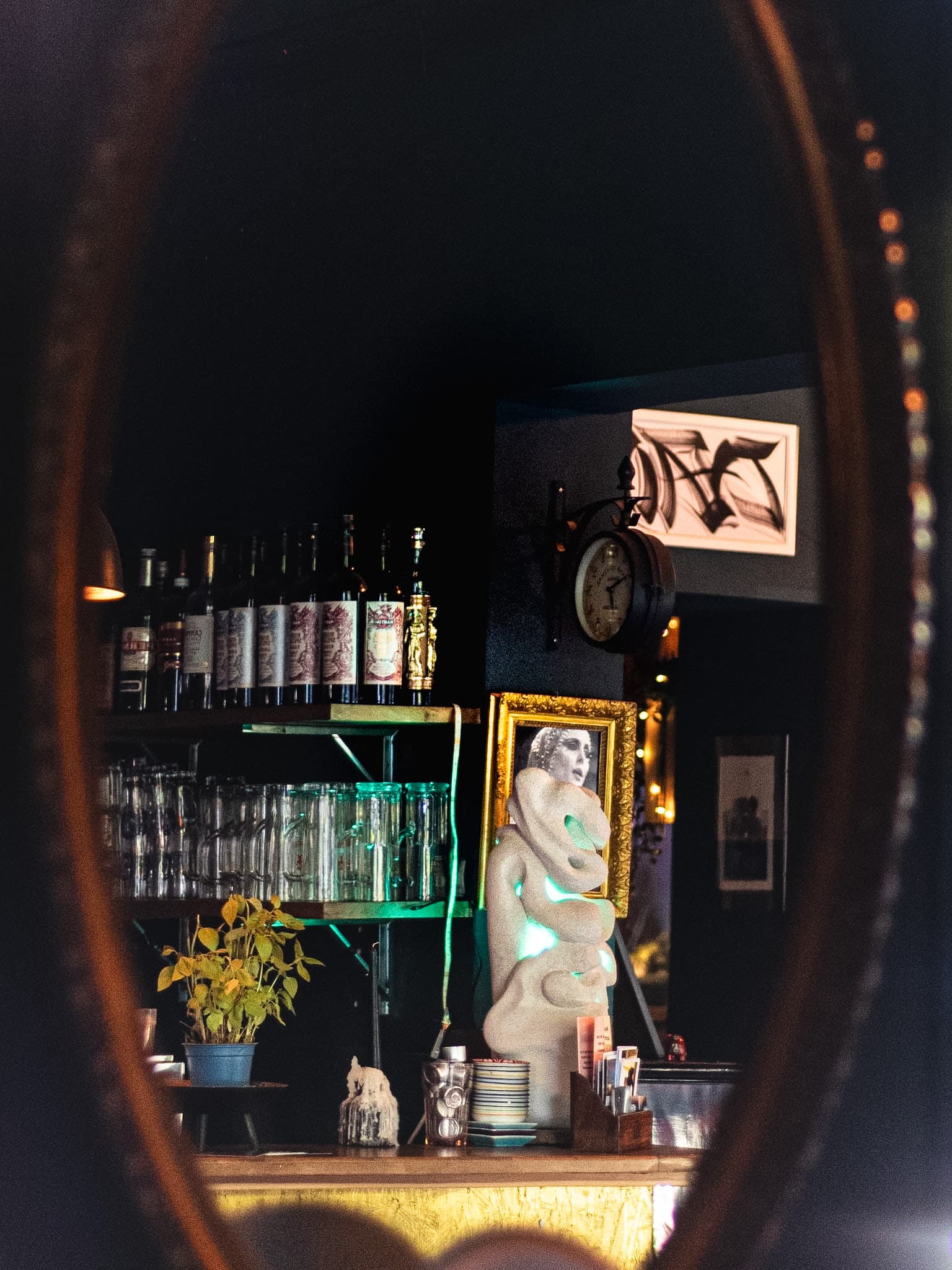 Atmospheric reflection of Beetroot Berlin bar counter, illuminated art sculpture, and botanical bottles through an antique oval mirror